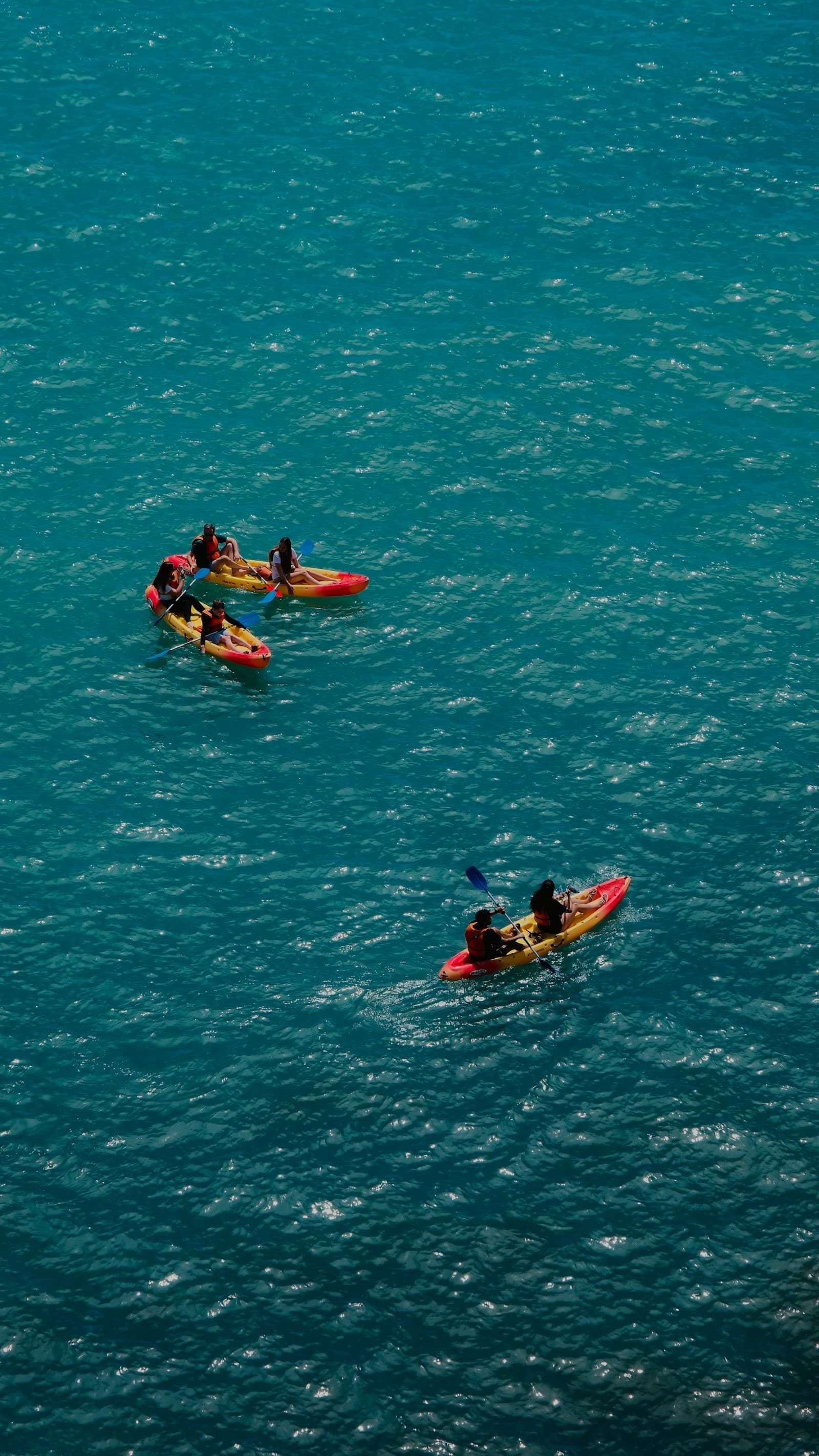 Guests paddling bright red and yellow kayaks across turquoise sea, seen from above